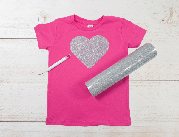 pink shirt with heart cut from glitter iron on, a Cricut weeding tool and a roll of glitter heat transfer vinyl